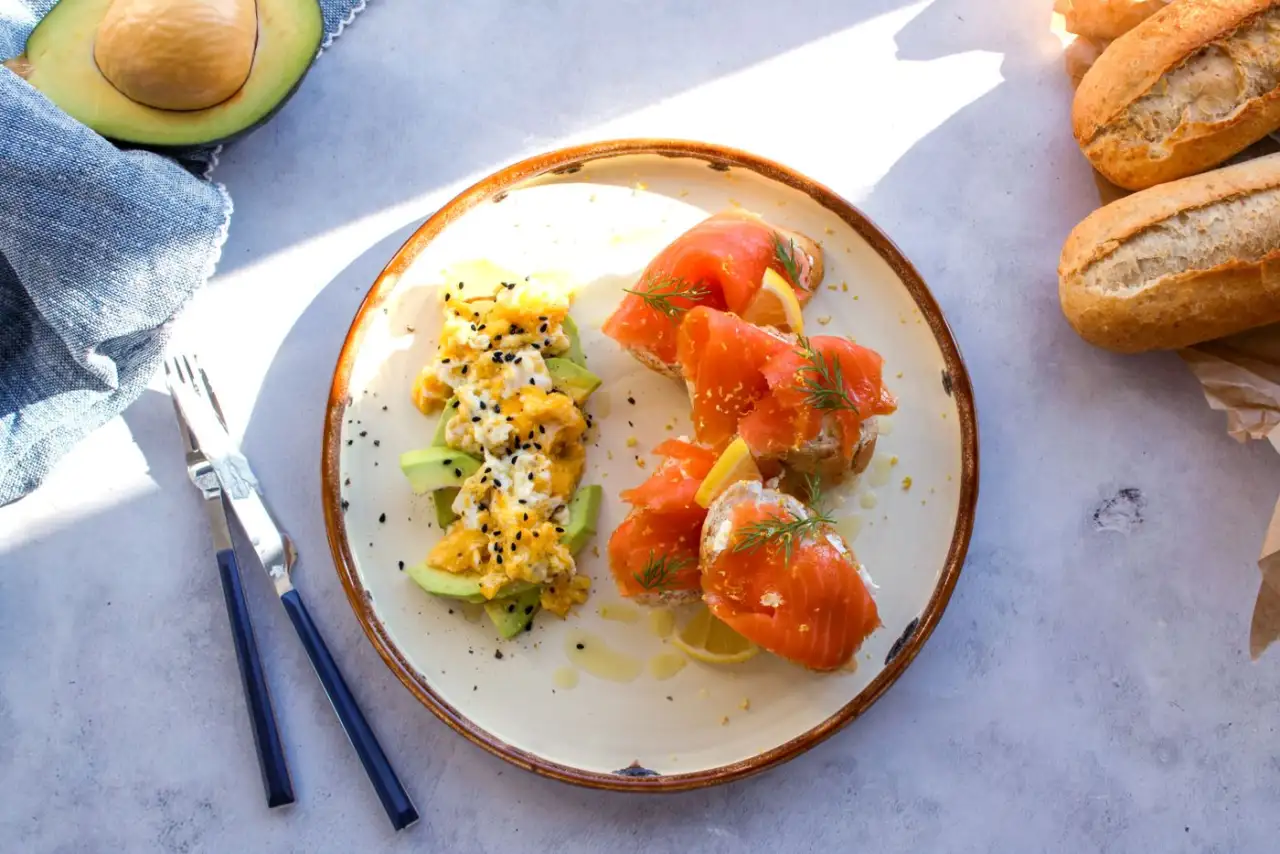 Frukosttallrik med rökt lax, äggröra och avokado bredvid färskt bröd
